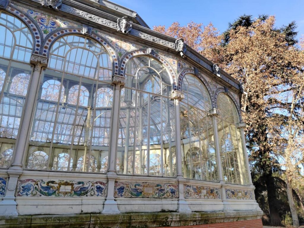 Iron and glass facade of the Crystal Palace in Madrid with white arches, decorative tiles and trees under a clear sky