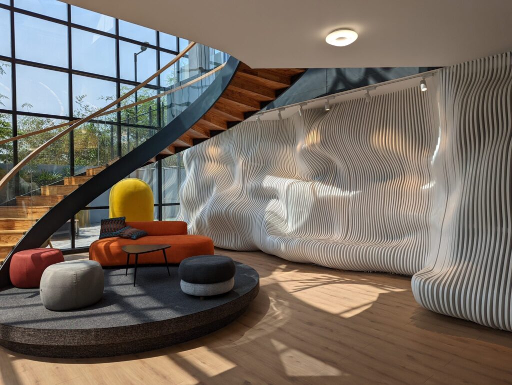 Modern lounge with a curved wooden staircase, a sculpted white wavy wall and colorful seating in sunlight from tall windows