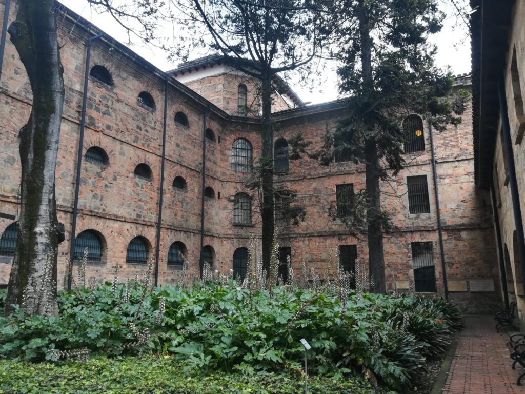 Red brick building with arched windows seen across a courtyard of tall trees and leafy plants