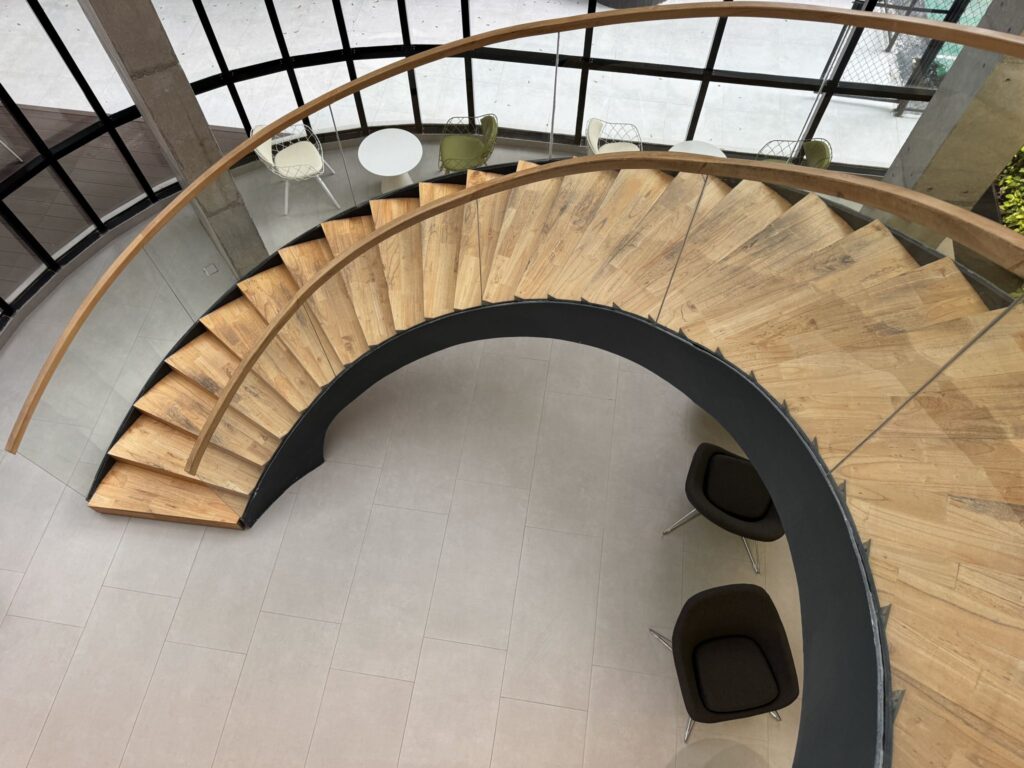 Spiral staircase with wooden steps and a glass railing curving down to a seating area with two black chairs